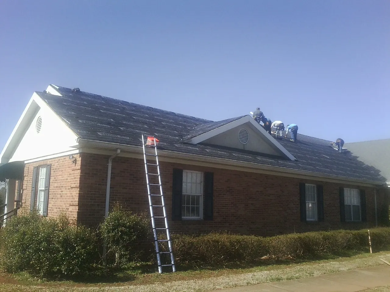 Crew tearing shingles off a brick ranch roof with a ladder set — Emergency Roof Repair work by our crew in Saint James