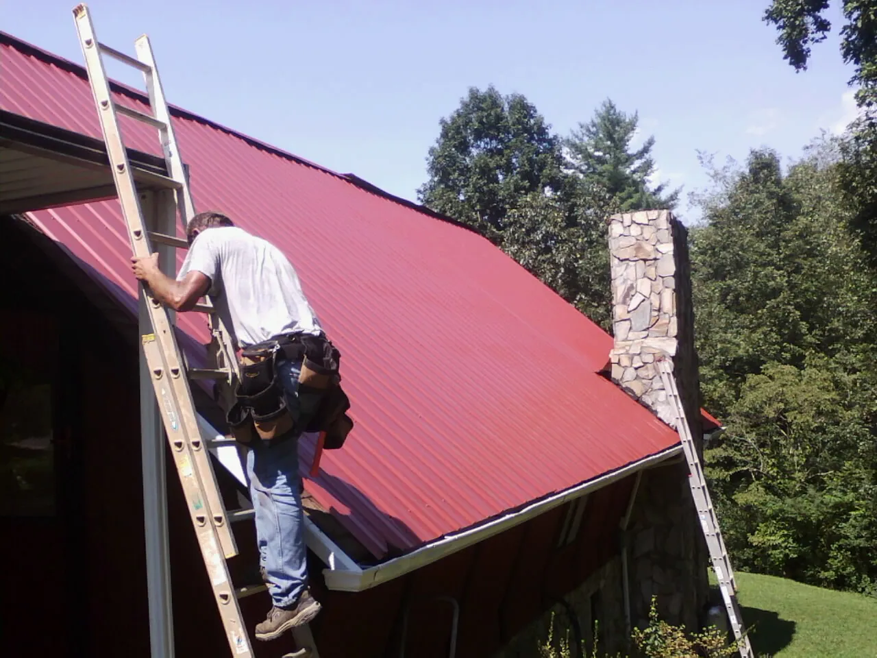 Roofer on a ladder beside a red metal roof and stone chimney — Soffit & Fascia Repair by Roofing Brothers in Saint James